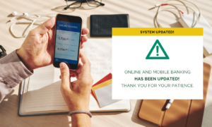 A pair of hands holding a phone with online banking on it, with a text box that reads: System Updated! Online and Mobile Banking has been updated! Thank you for your patience.