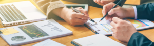 Cover photo - hands with pencils and pens pointing at graphs and charts on table